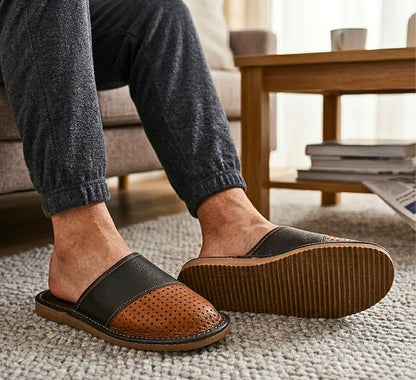 A person wearing brown and black slip-on mule slippers with perforated detailing, shown in a cozy living room setting with a sofa and wooden table in the background.
